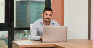 A male behind a laptop at a desk in front of a window